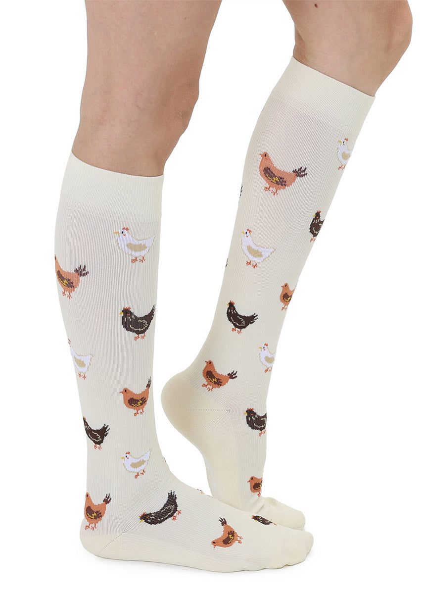 Off-white knee-high compression socks with a repeating chicken pattern worn by a person in front of a white background.