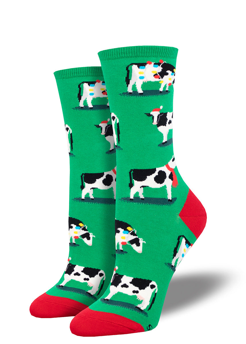 Green holiday socks for women with a red heel and toe featuring an allover pattern of black and white cows wearing Santa hats, scarves, and Christmas lights. 