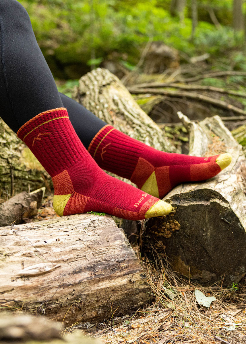A model wearing deep red wool hiking socks poses sitting outside with their feet on top of a log.
