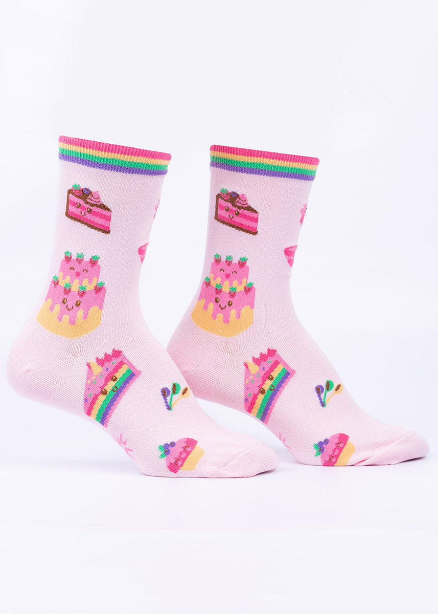 Pink women's crew socks with rainbow-striped cuffs feature a pattern of smiling cakes with rainbow fillings, pink and chocolate icing and various decorations.