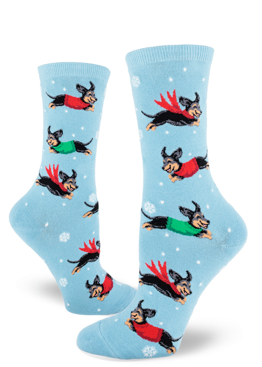 Light blue crew socks for women show a repeating pattern of snowflakes and dachshunds wearing red and green sweaters and scarves playing in the snow.