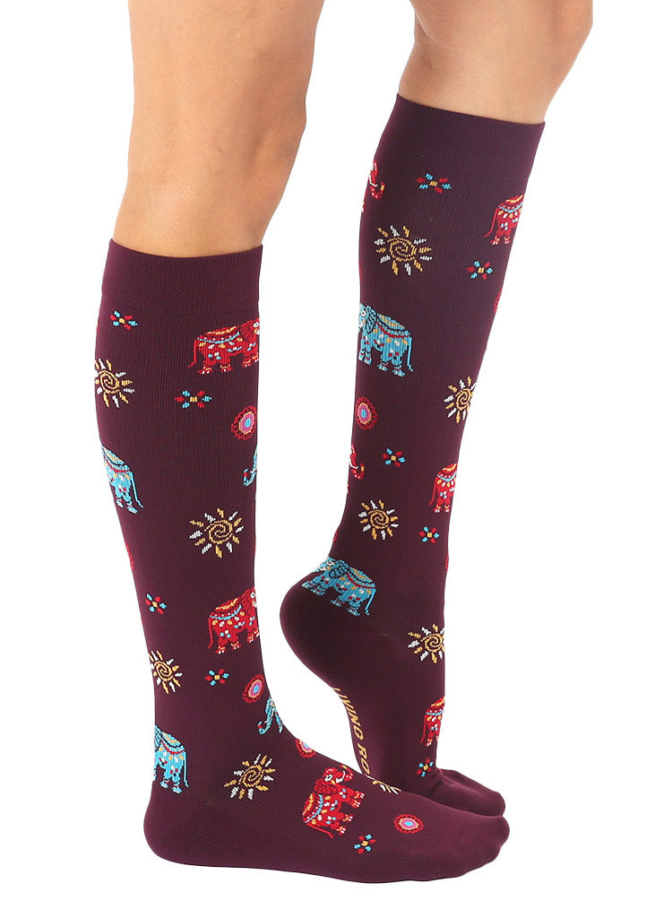 Side view of maroon knee-high compression socks with an allover pattern of red and blue painted elephants plus abstract sun designs.