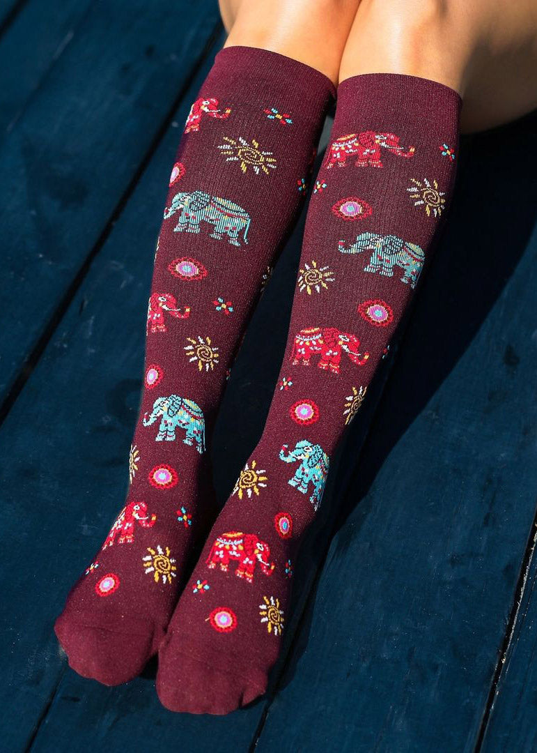 A model wearing elephant-themed compression knee socks poses sitting on a dark blue couch.