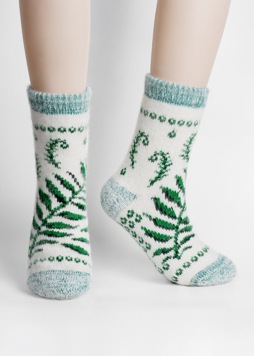 White socks with green fern and fiddlehead patterns worn by a person standing in front of a light gray backdrop.