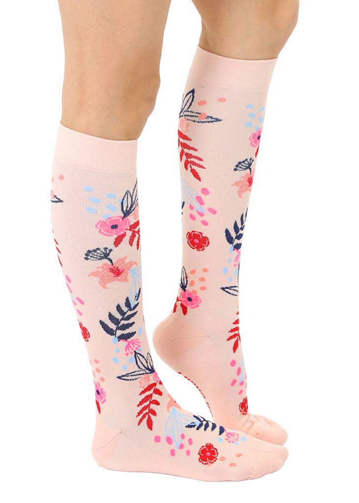 A side view of light pink knee-high compression socks with an allover pattern of red, pink, navy, and light blue floral motifs.