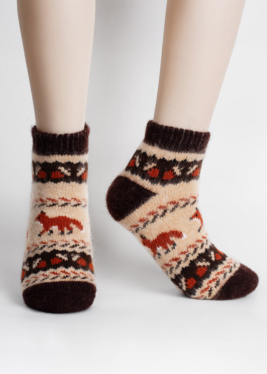 Fuzzy brown and tan wool ankle socks with red-orange fox and acorn patterns worn by a person standing in front of a light gray backdrop.