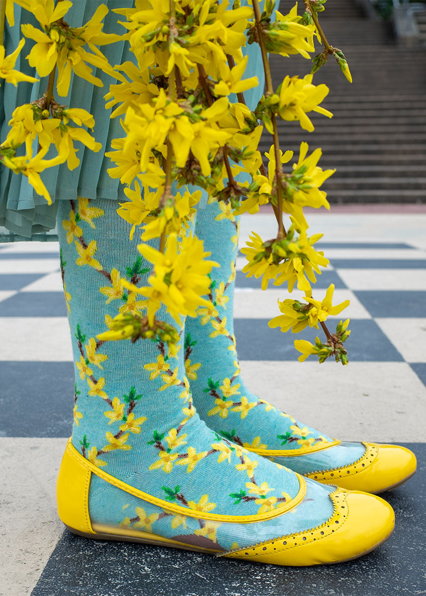 Floral knee-high socks feature a repeating pattern of forsythia flowers, the yellow buds arranged along brown branches, over a pale aqua heather background.