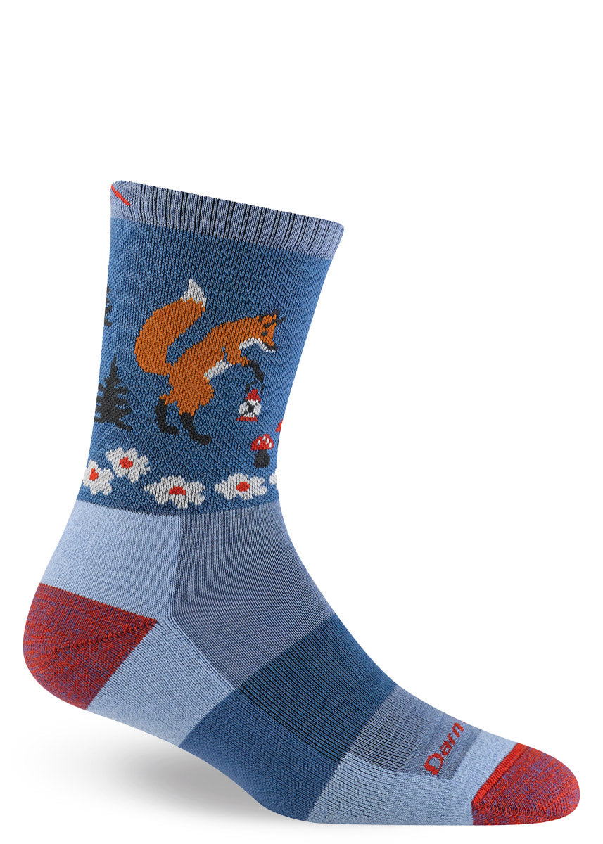 Blue hiking crew socks with red accents depict a whimsical scene of a fox carrying a lantern through a forest full of trees, flowers and small red mushrooms with a crescent moon up above.
