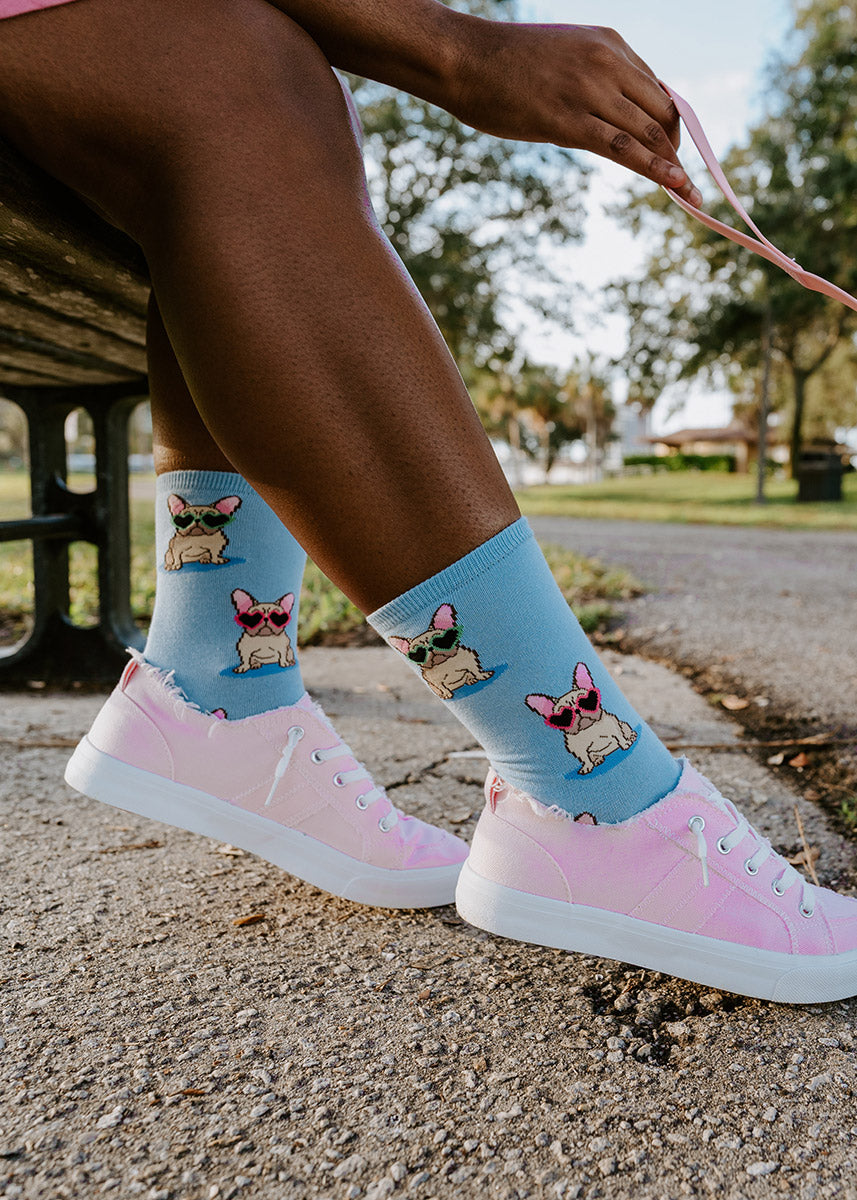 A model wearing novelty socks featuring french bulldogs wearing heart-shaped sunglasses poses holding a dog leash and sitting on a park bench.