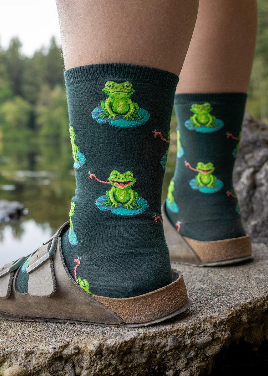 A female model poses wearing dark green novelty socks for women with an allover pattern of green frogs sitting on lily pads wearing brown sandals and standing outside on a rock.