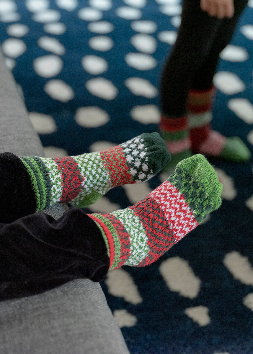 A toddler poses sitting on a couch wearing intentionally mismatched holiday socks with different sections featuring various geometric patterns like stripes and dots in shades of red, green, and white.