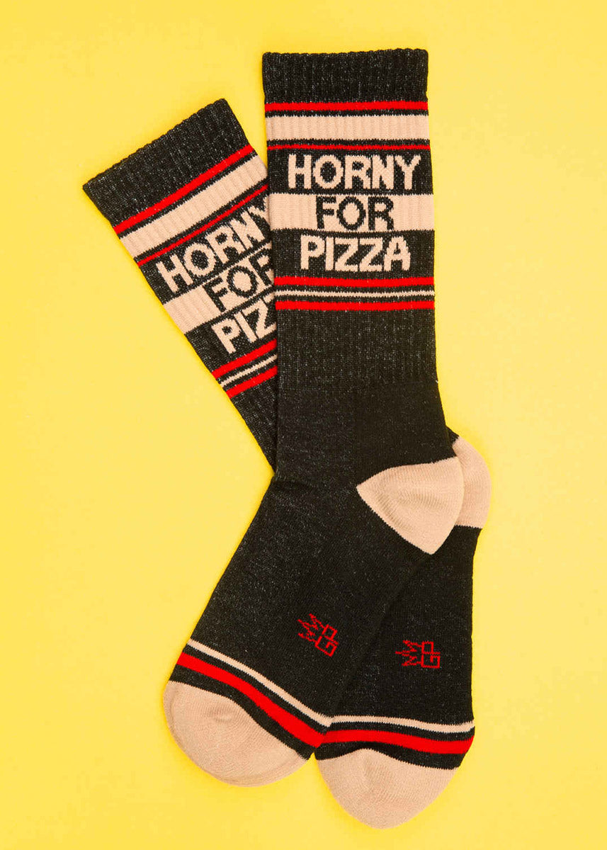 Retro-style unisex gym socks that say &quot;HORNY FOR PIZZA&quot; on a black background with bright red and tan accents lay flat against a bright yellow background.