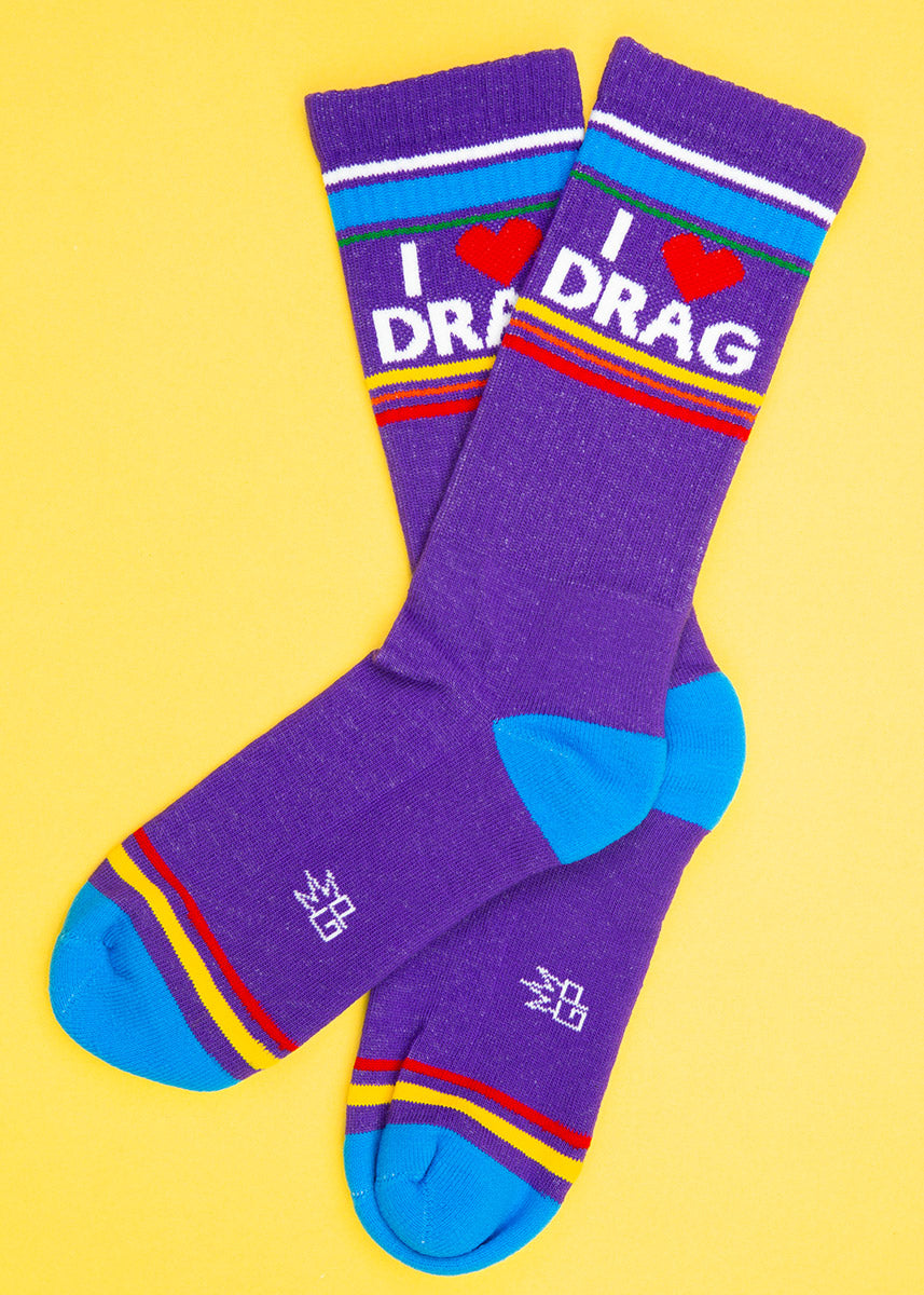 A pair of purple gym-style novelty socks that read &quot;I ❤️ Drag&quot; are laid out flat against a pastel yellow background.