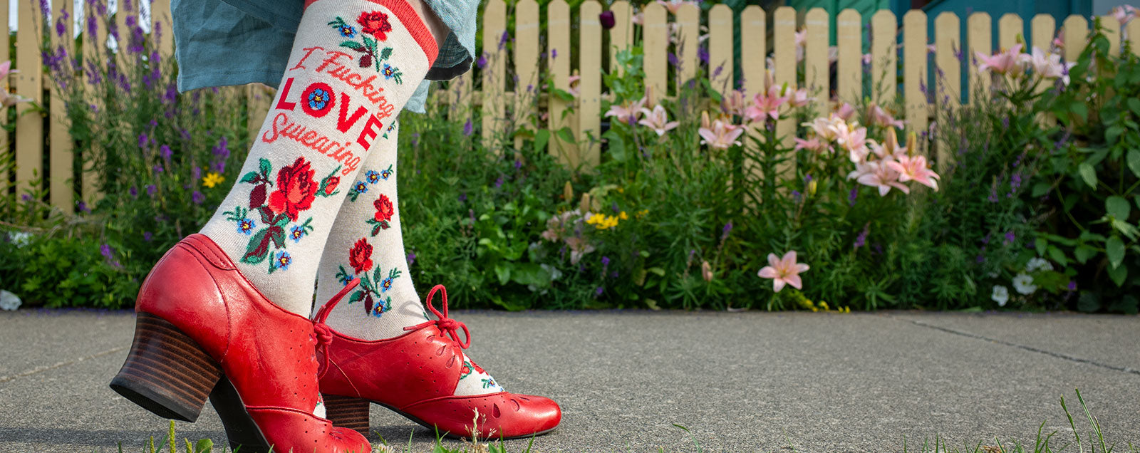 A woman wears cream floral crew socks that say “I Fucking LOVE Swearing" on the leg while walking next to a picket fence and garden.