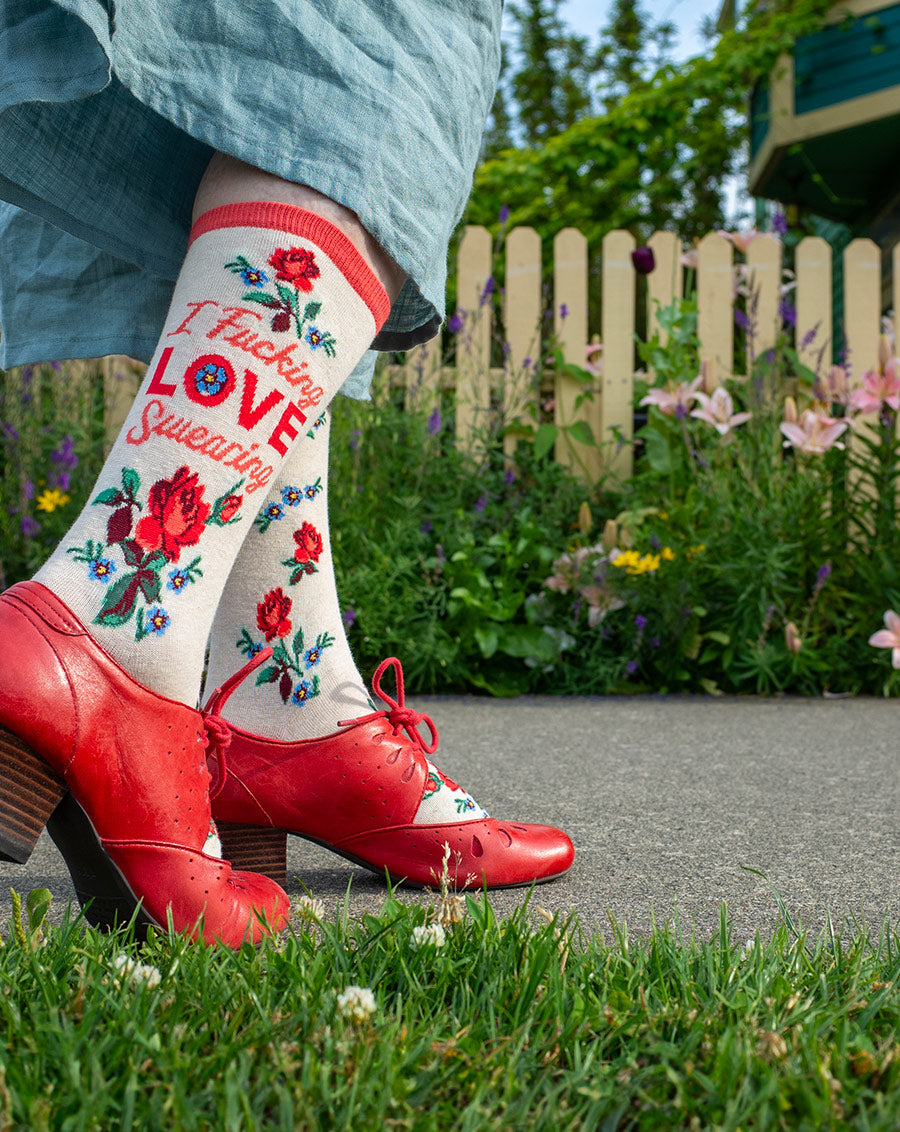 A woman wears cream floral crew socks that say “I Fucking LOVE Swearing" on the leg while walking next to a picket fence and garden.