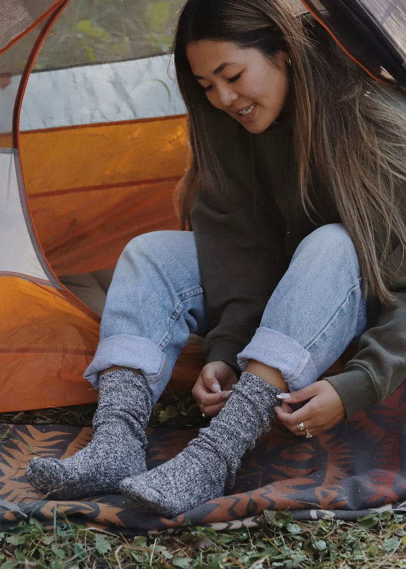 A model wearing black and white slouchy knit socks poses in a camping tent outside.