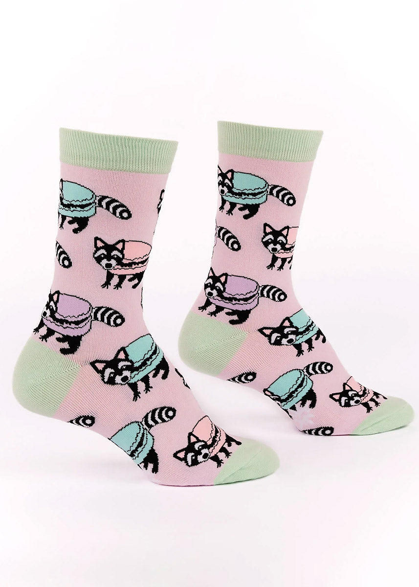 Light pink crew socks for women with an allover pattern of black and white raccoons with colorful pastel macarons as their bodies.