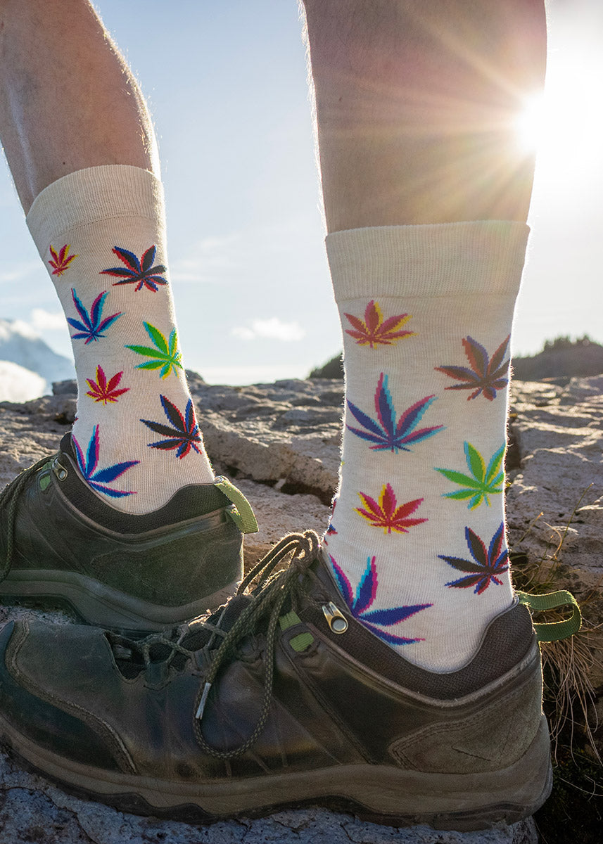 A male model wearing crew socks that feature colorful weed leaves against a cream background and hiking sneakers poses standing on a rock.