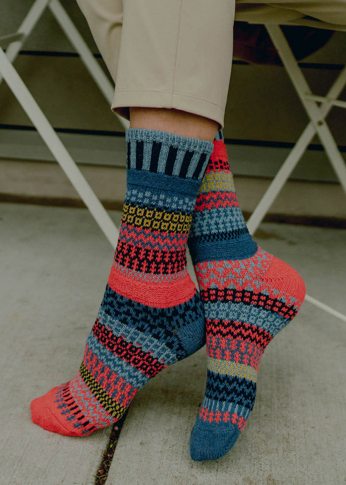 A model wearing mismatched socks in shades of blue, coral, and moss green poses on the sidewalk.