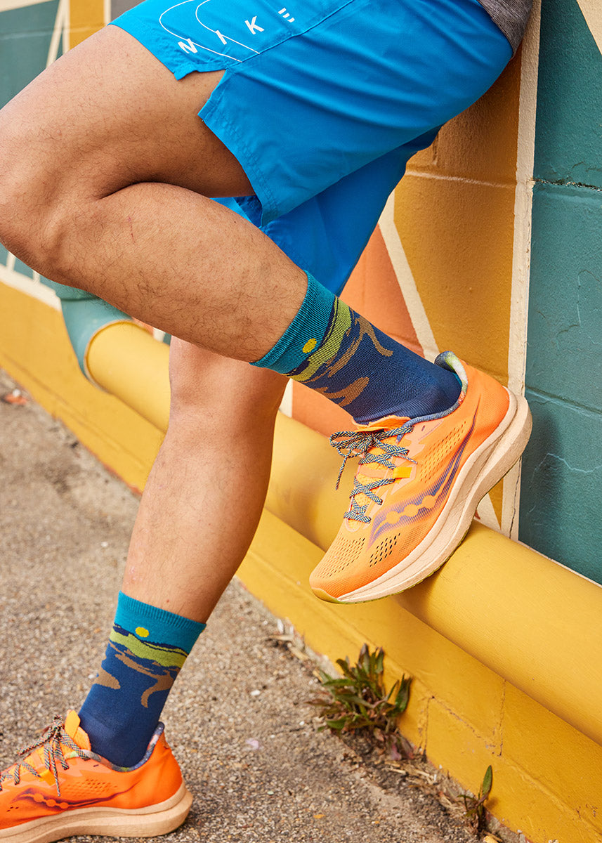 Dark blue lightweight hiking socks for men with a green and brown mountain landscape against a teal sky with a yellow sun. 