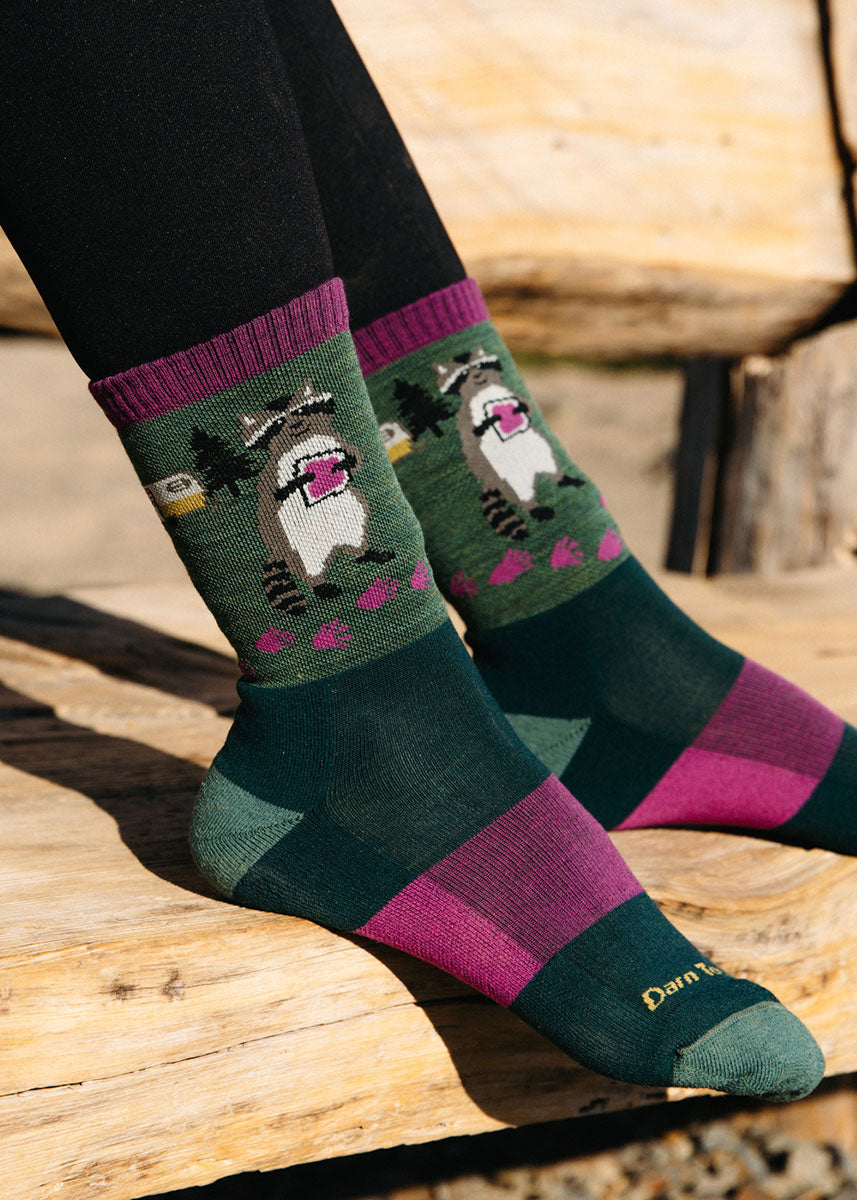 A model wearing green and pink striped hiking socks with a cartoon raccoon design poses resting their feet on a plank of wood.