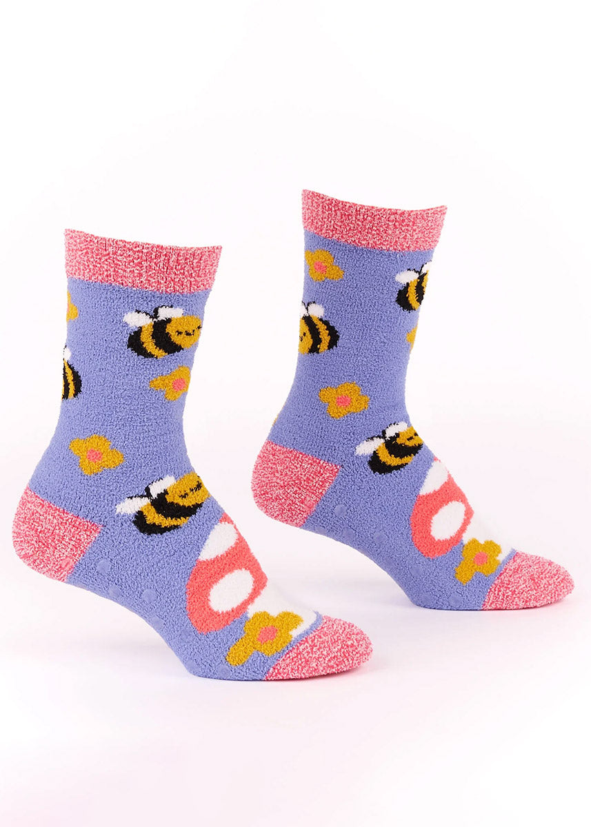 Purple fuzzy crew socks for women with pink accents and an allover pattern of smiling bumblebees, flowers, and a large toadstool mushroom.