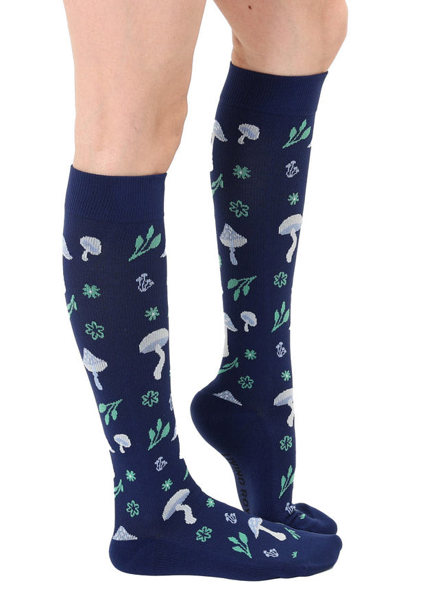 A female model poses to the side wearing navy knee-high compression socks with an allover pattern of white and light blue toadstool mushrooms and green foliage.