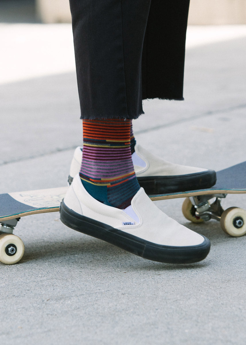 A model wearing navy, purple, and orange striped wool socks and white Vans poses on a skateboard.