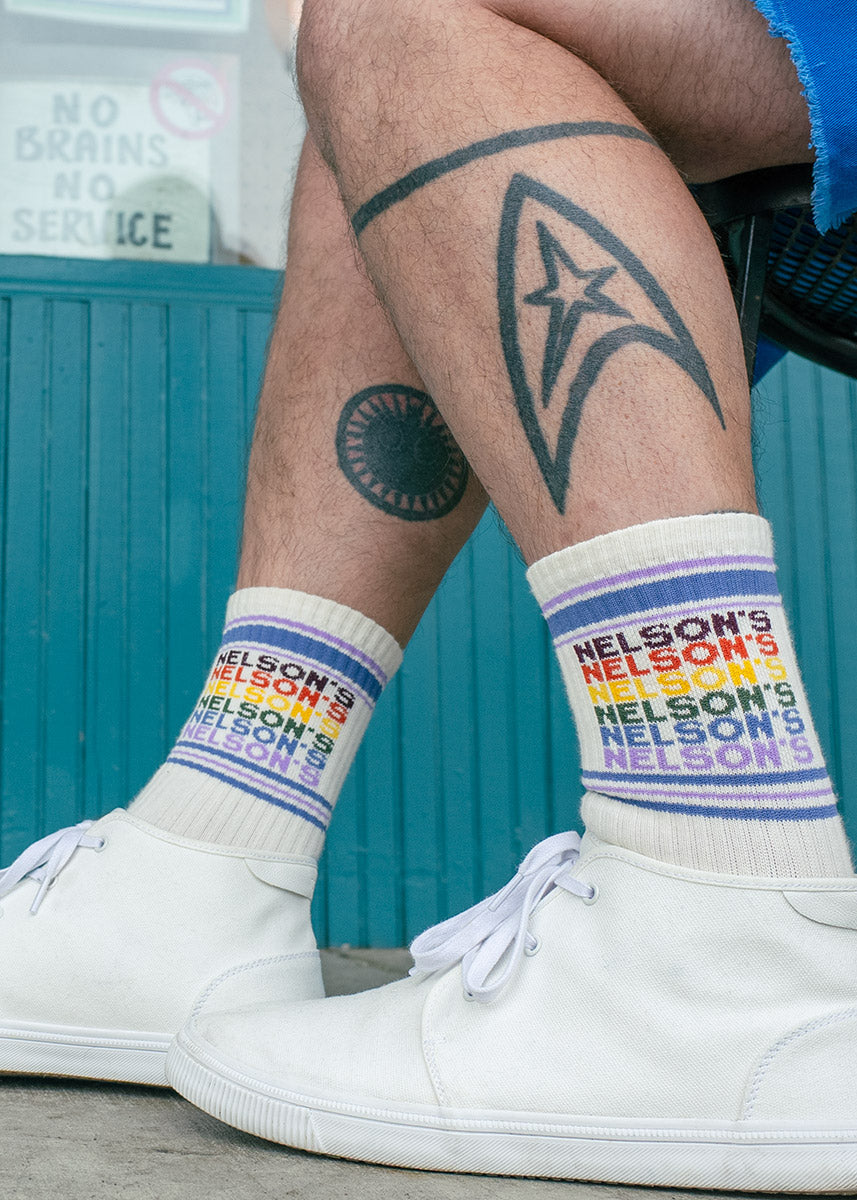 A Nelson&#39;s Market employee wears a pair of cream retro-style gym socks with vintage-inspired rainbow lettering spelling out “NELSON&#39;S&quot; while standing in front of the iconic Bellingham corner store and diner.