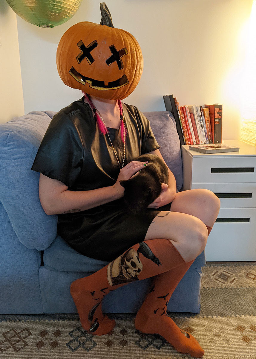 A model wearing knee socks inspired by Edgar Allen Poe featuring ravens, human skulls, antique books and feather pens poses sitting with a black cat in their lap and a jack-o-lantern over their head.