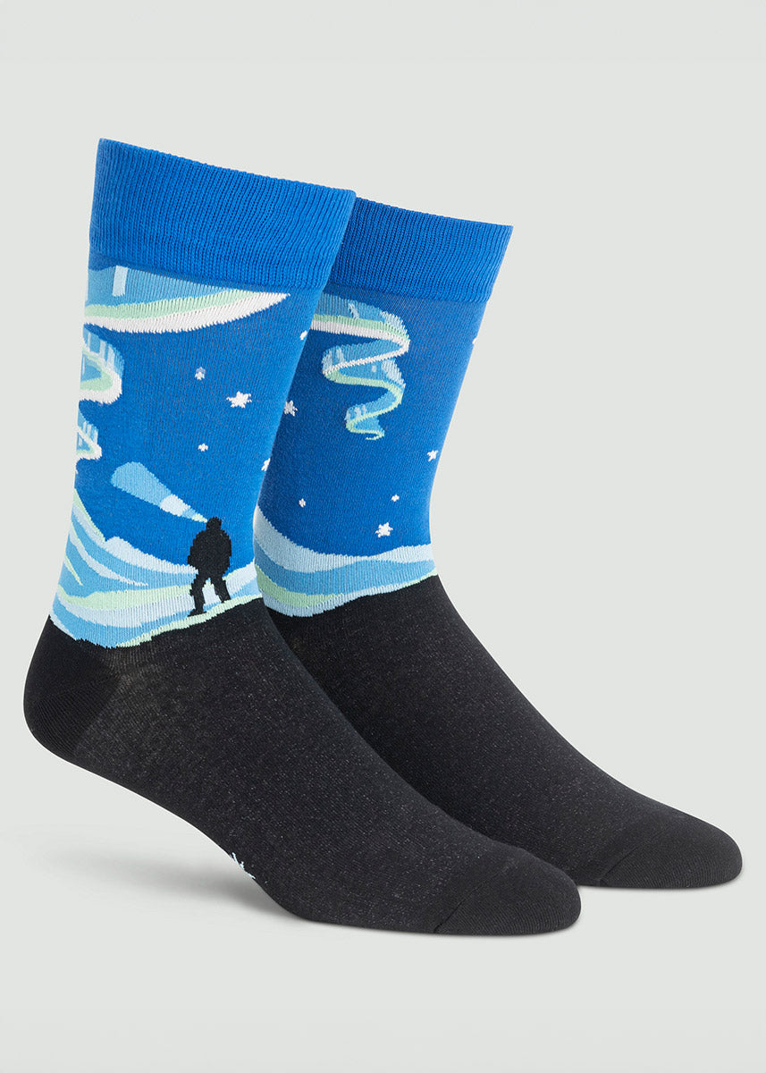 Glow-in-the-dark men&#39;s crew socks with the silhouette of a person taking in a view of the northern lights, with glowing stars and streaks of white against a dark blue sky background.