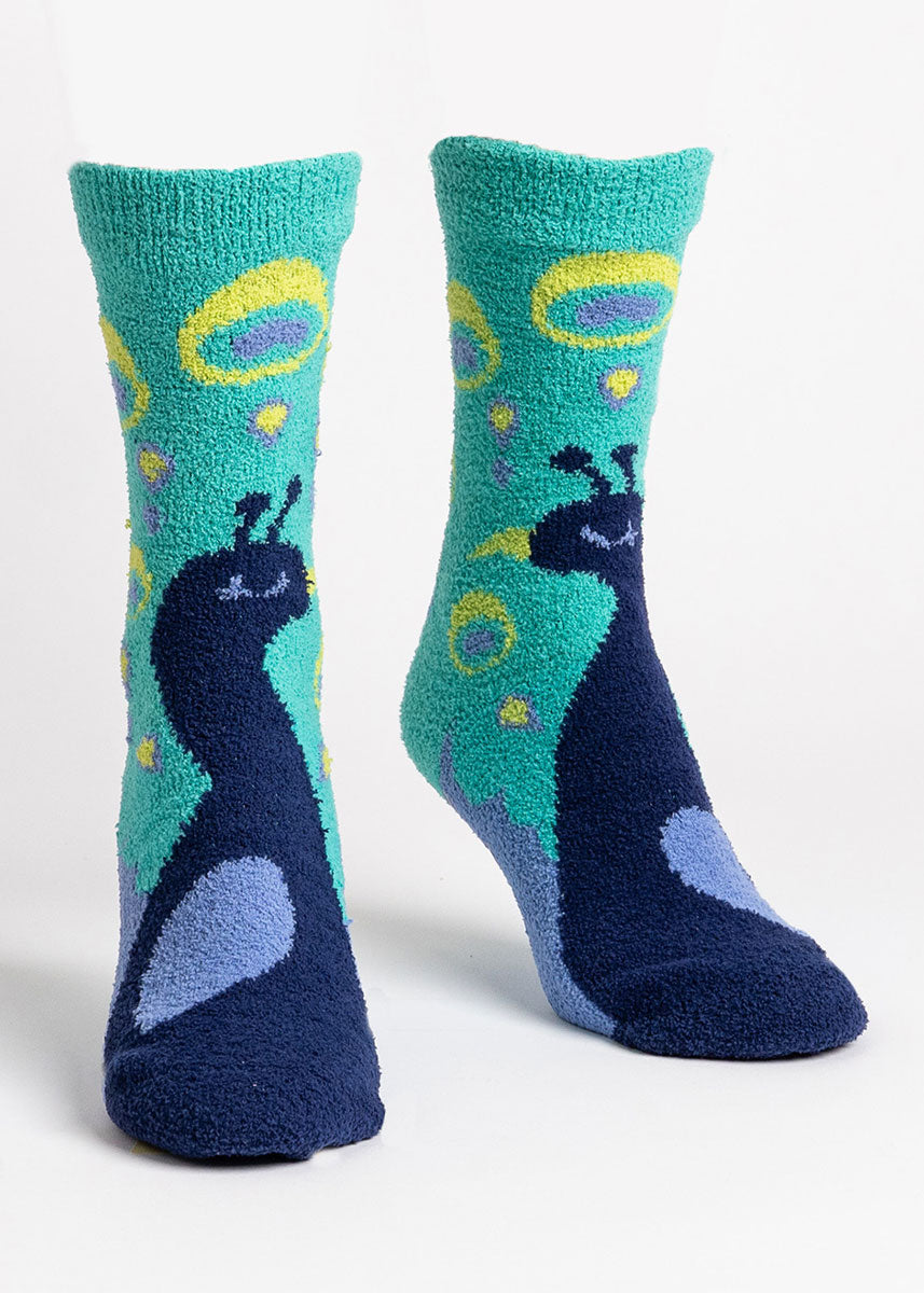 Fuzzy slipper socks for women that feature a dark blue peacock on each foot against a teal background. 