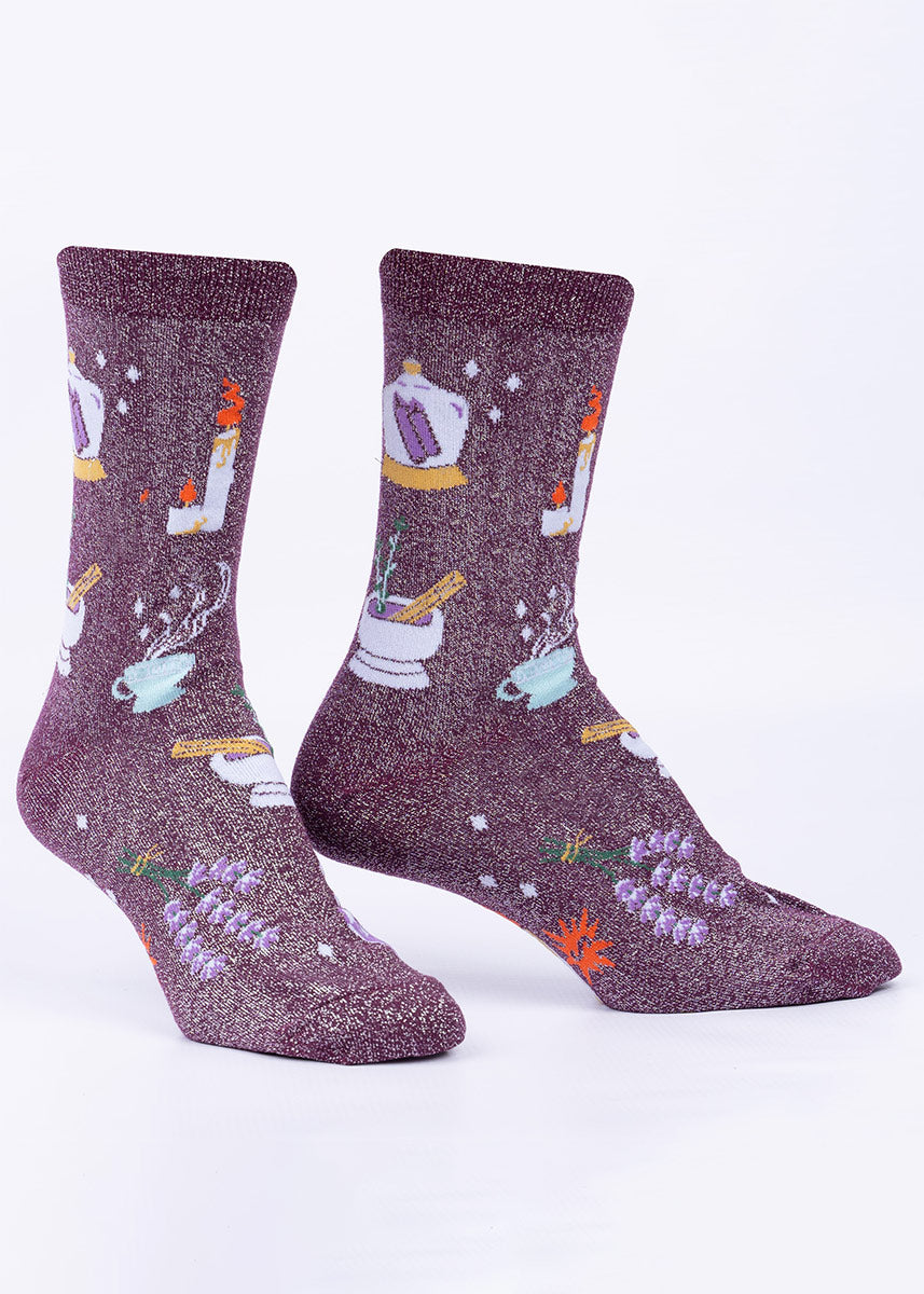 Crew socks with a shimmering purple background feature a witchy design with crystals, candles, lavender bundles and mortar and pestles.
