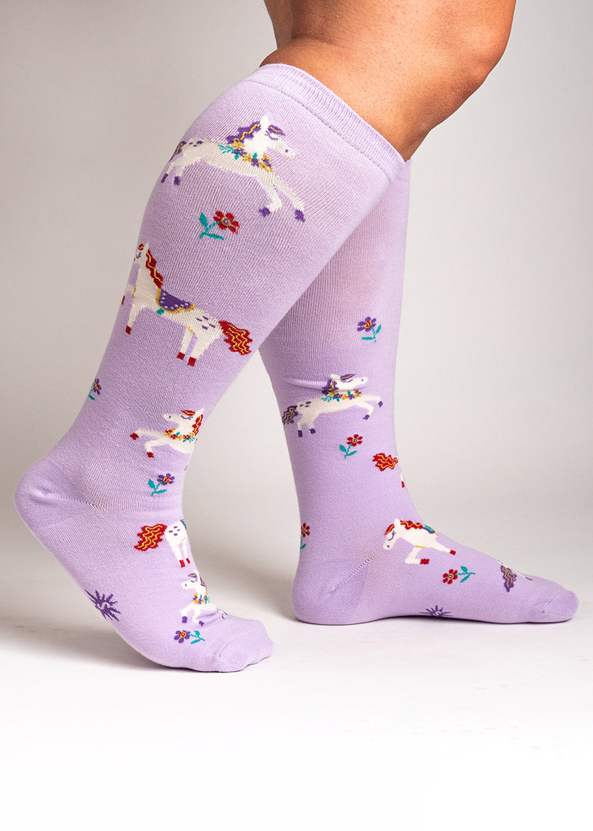 Lavender knee socks designed for wide calves with an allover pattern of white horses with colorful manes and accessories like floral necklaces and saddles, as well as flowers. 