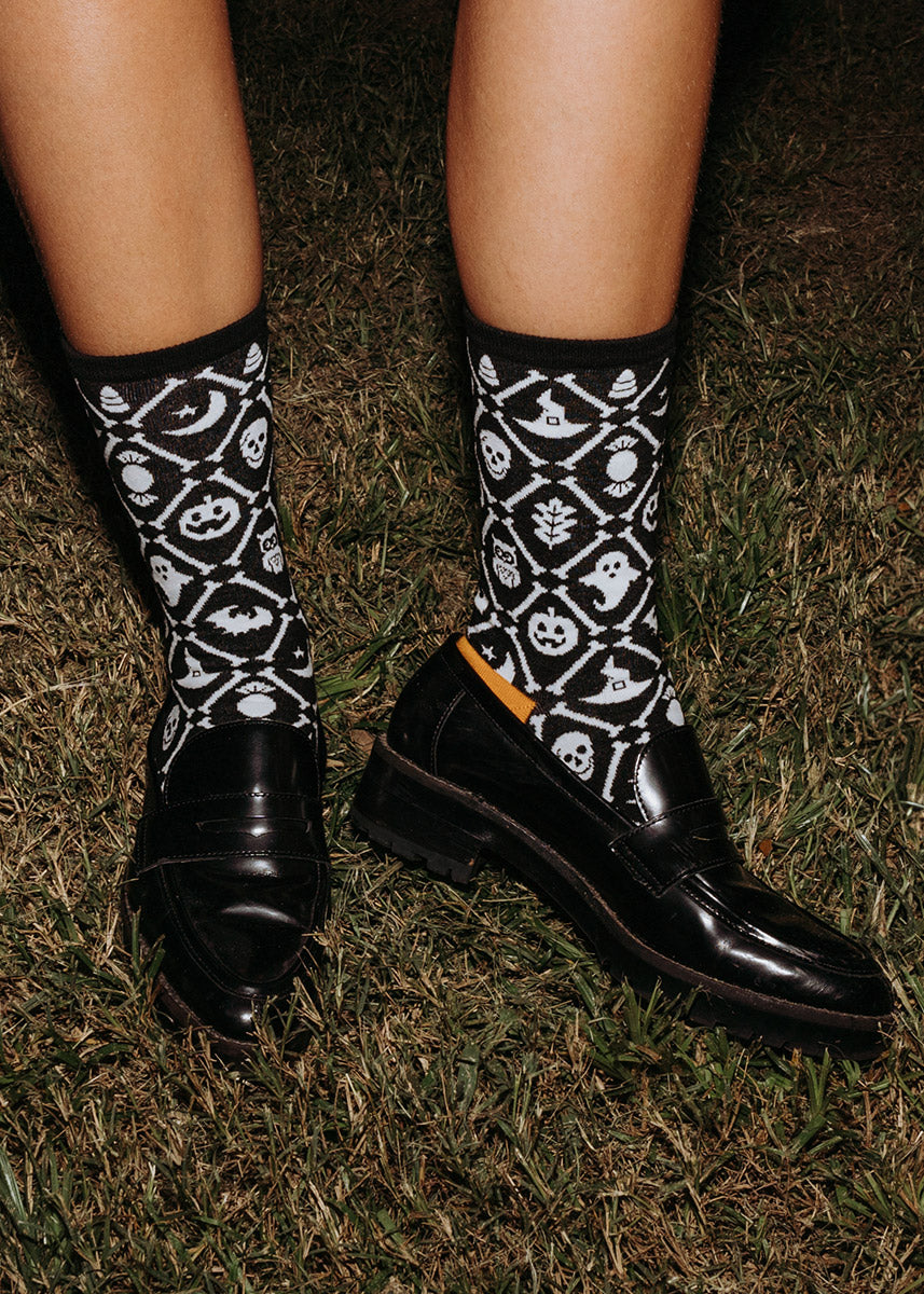 A model wearing Halloween-patterned novelty socks and black loafers poses outside in the grass at night.