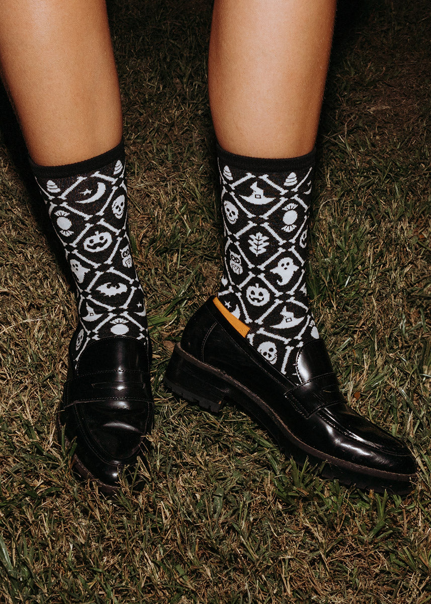 Black and white women's crew socks with orange accents feature a repeating pattern of Halloween icons arranged within a diamond pattern bordered with bones.