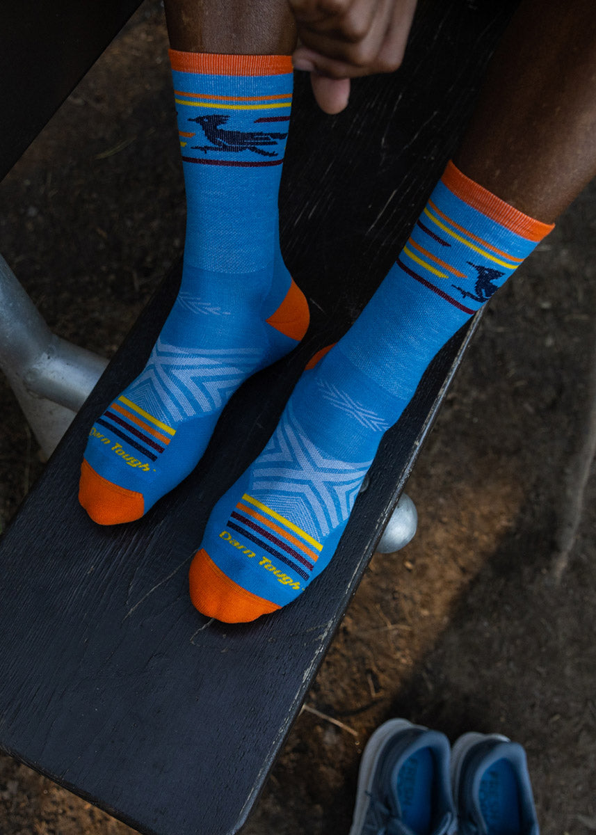 A model wearing blue roadrunner-themed wool socks poses sitting on an outdoor bench.