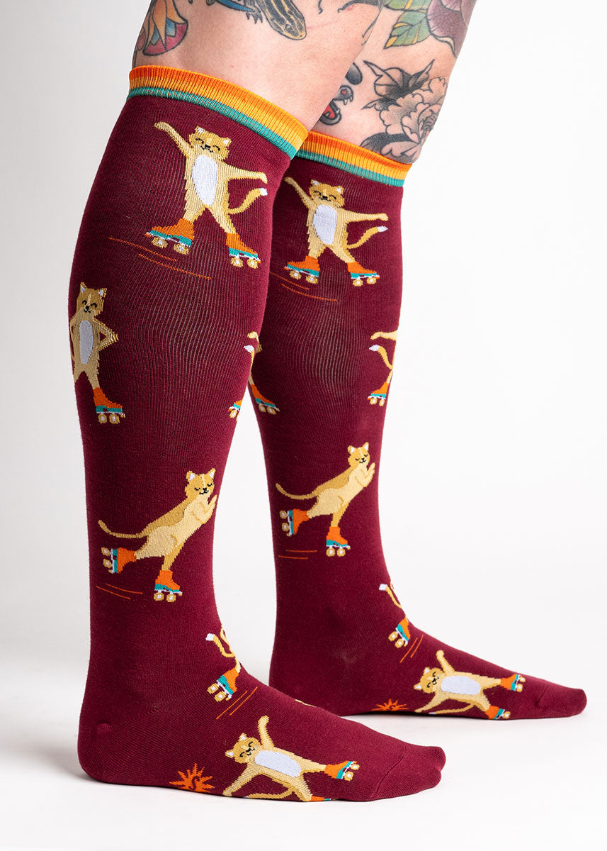 A model poses wearing red knee-high socks for women that feature an allover pattern of yellow cats wearing roller skates and skating in different poses.