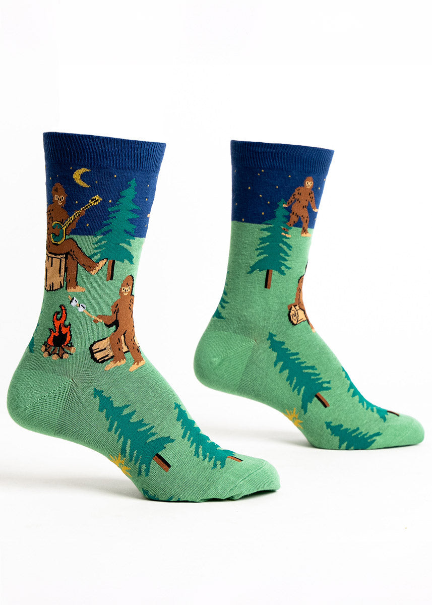 Green and navy crew socks for women that feature a nighttime forest scene of multiple Sasquatches sitting around a campfire, roasting marshmallows and playing the banjo. 