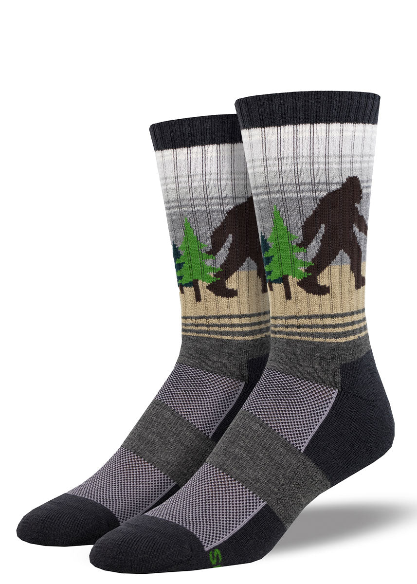 Black, gray, and light gray gradient striped hiking socks for men that depict a black Sasquatch silhouette in a green pine tree forest.