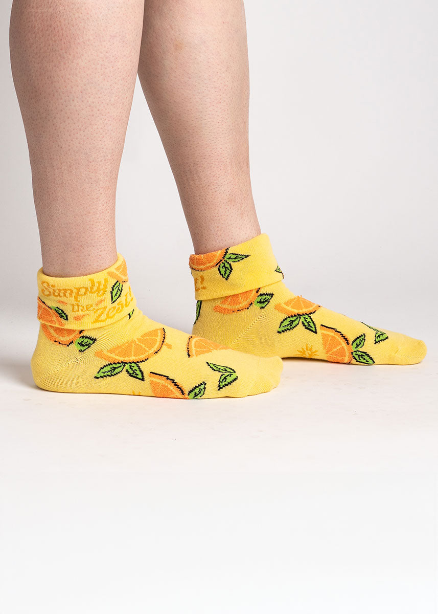 A model poses against a white background wearing yellow turn-cuff ankle socks for women with an allover pattern of fuzzy orange slices and the words "Simply the Zest" written on the cuff.