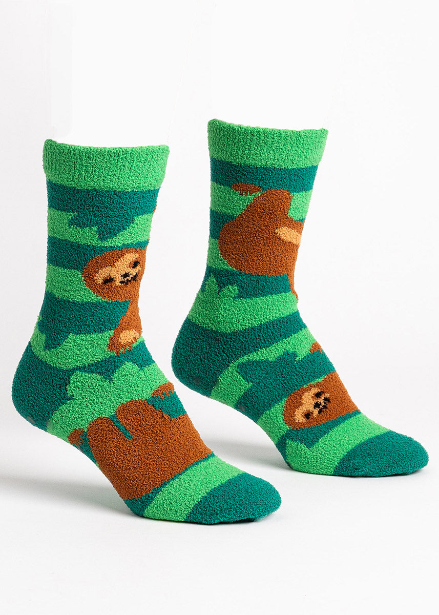 Fuzzy green slipper socks for women that feature a green leafy striped design with brown smiling sloths hanging from the stripes like trees.