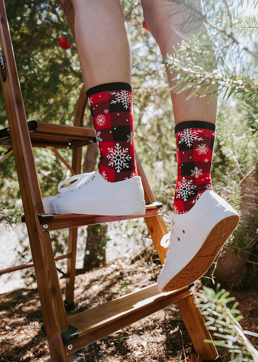 A model wearing red and black plaid and snowflake crew socks and white sneakers poses climbing a ladder in the forest.