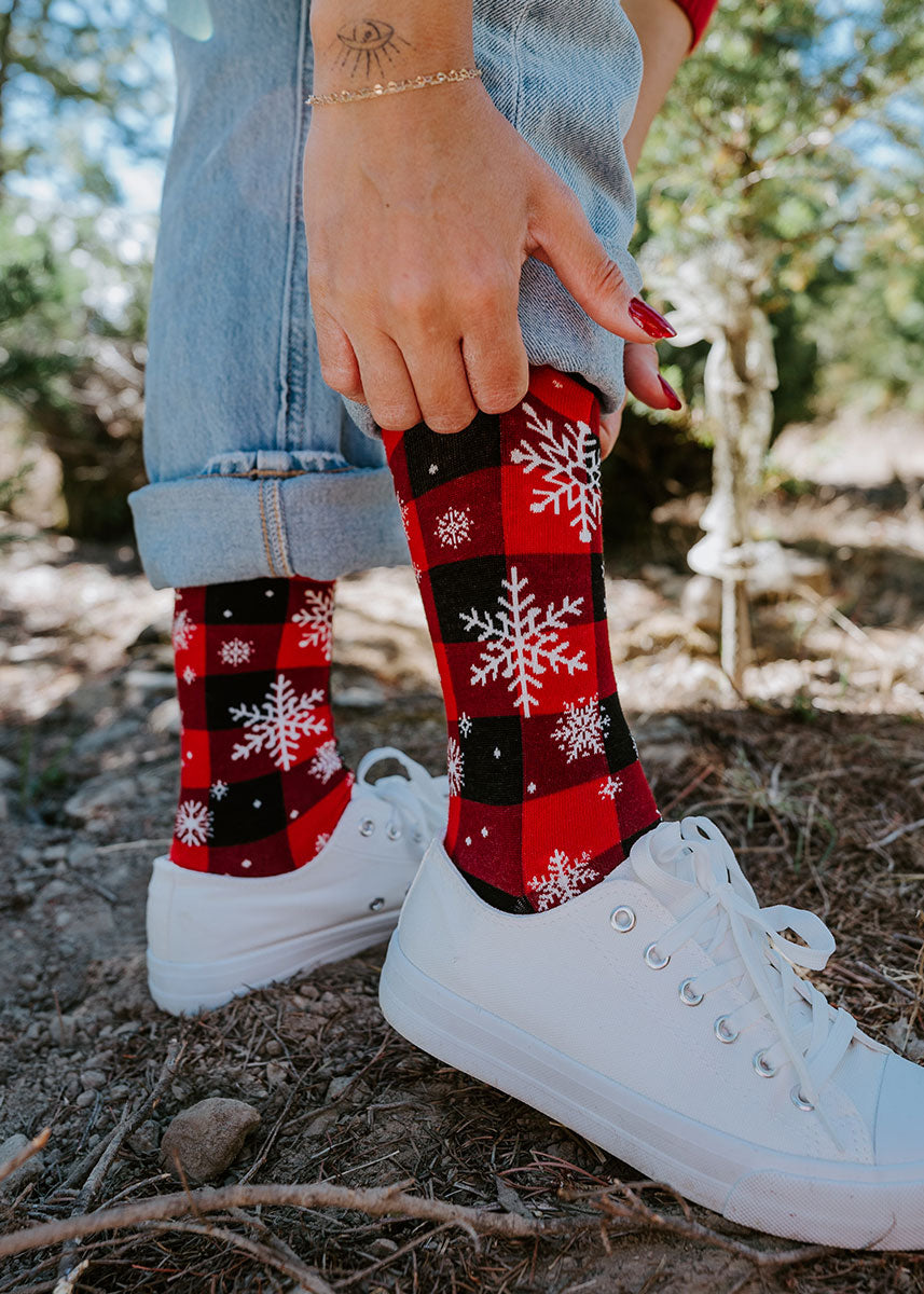 A model wearing red and black plaid and snowflake crew socks and white sneakers poses in the forest pulling up the cuff of their jeans.