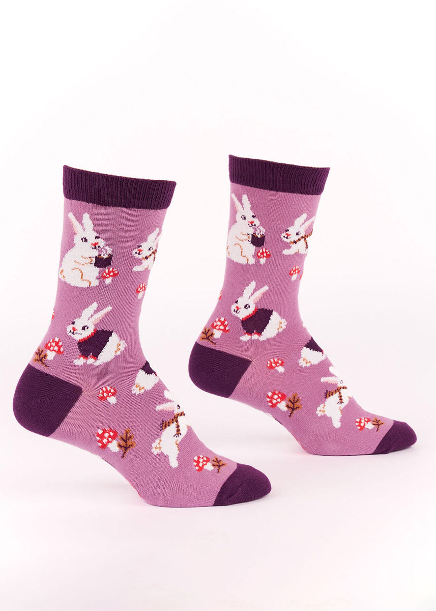 Purple crew socks for women with an allover pattern of fuzzy white bunnies  dressed in warm clothes, as well as fall foliage such as red toadstool mushrooms and leaves.