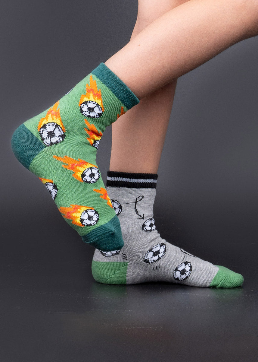 A child model poses wearing mismatched soccer-themed socks, with one green sock featuring flaming soccer balls and one gray sock with soccer balls and arrows.