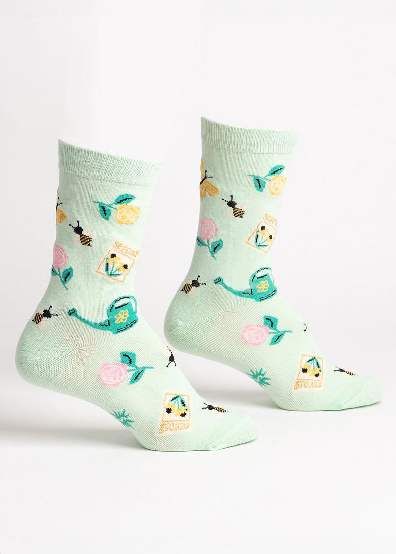 Mint green crew socks for women with an allover pattern of yellow and pink roses, yellow butterflies, bumblebees, seed packets, and green watering cans.