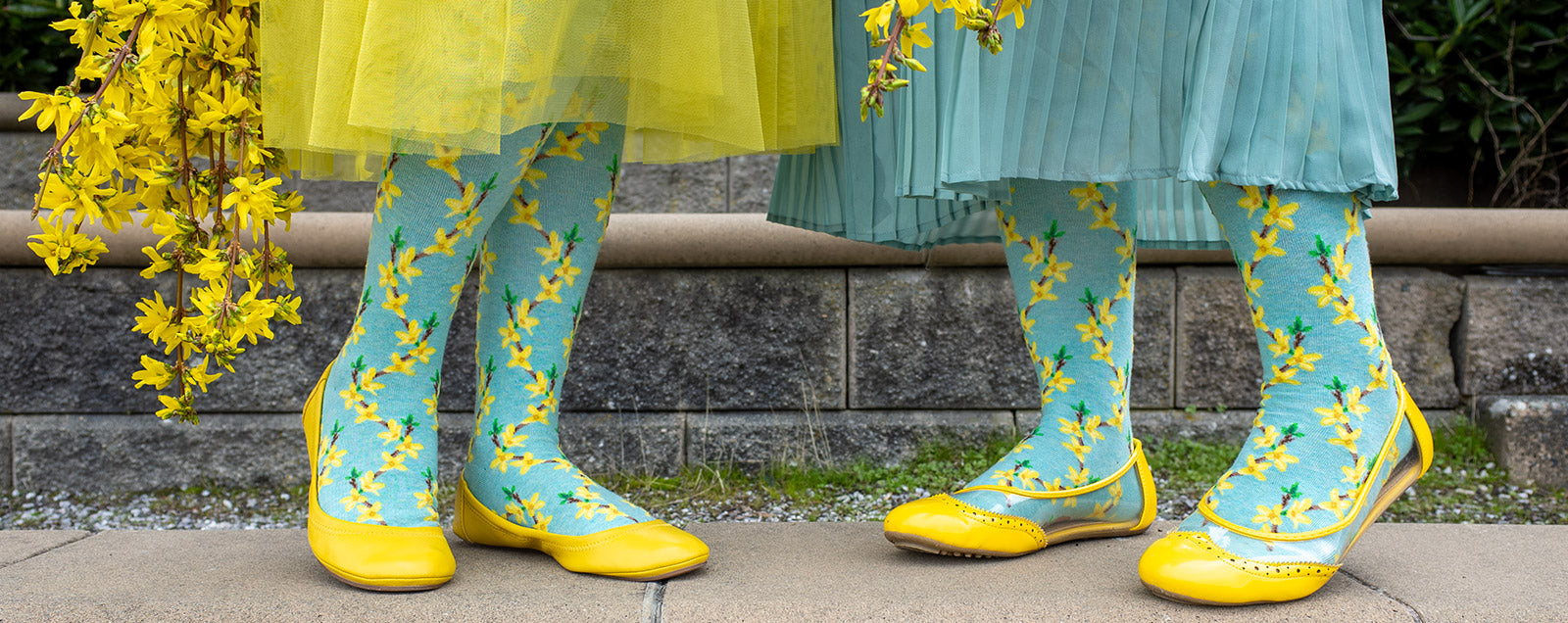 Two models wear yellow ballet flats and floral knee socks with bright yellow forsythia flowers over a pale aqua background.