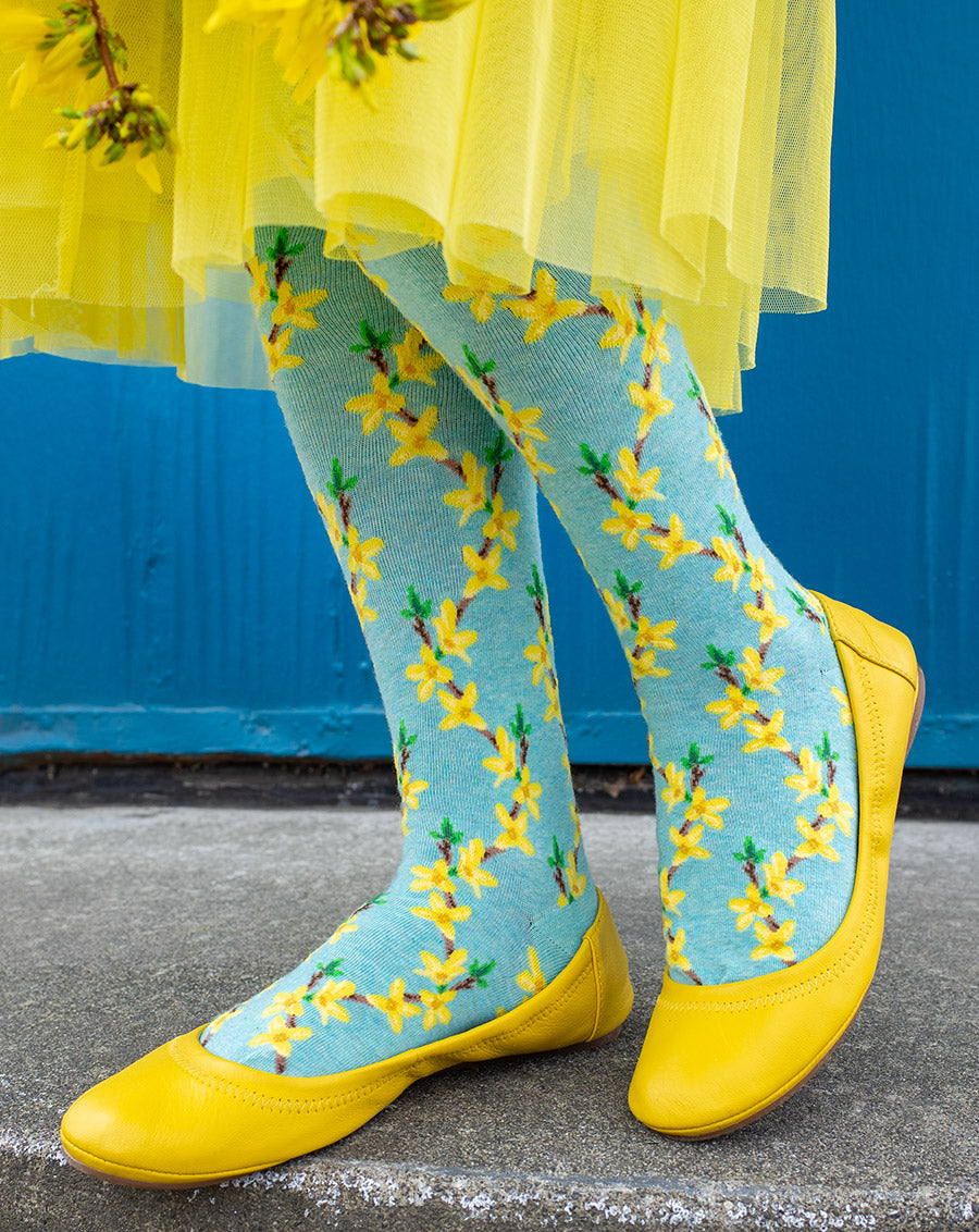 A model wears yellow ballet flats and floral knee socks with bright yellow forsythia flowers over a pale aqua background.