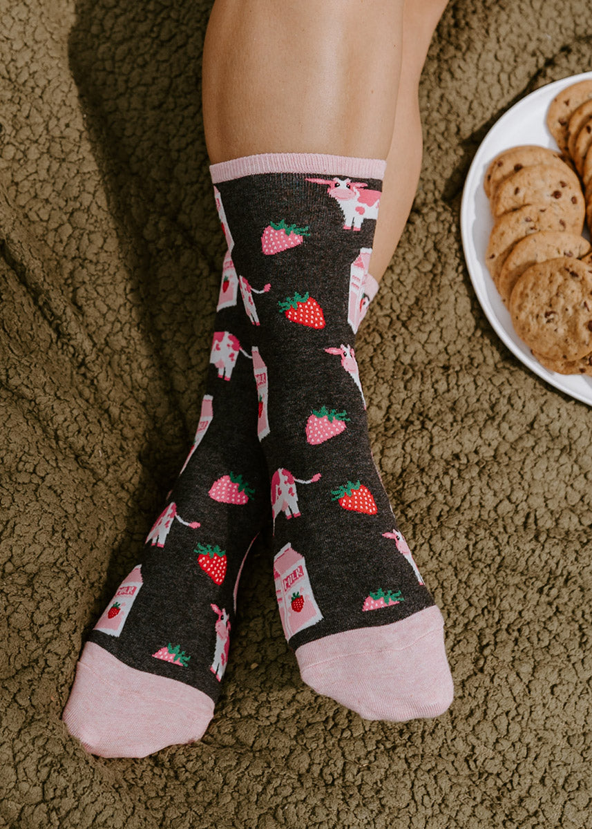 A model wearing strawberry milk and cow-themed novelty socks poses next to a plate of cookies.